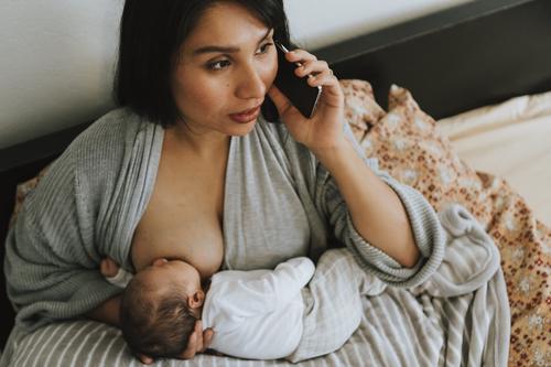 Baby latched on, laying on breastfeeding pillow, with parent hunched over supporting baby’s neck with one hand and talking on the phone with the other 