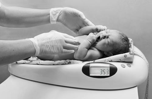 Black and white of a baby on an infant scale with adult gloved hands that have just placed them there 